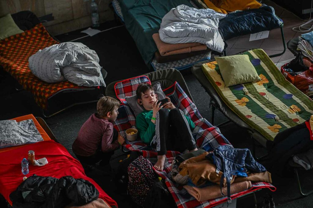 Two boys are in a shelter. One of them lies on a cot looking at his phone while the other looks on.