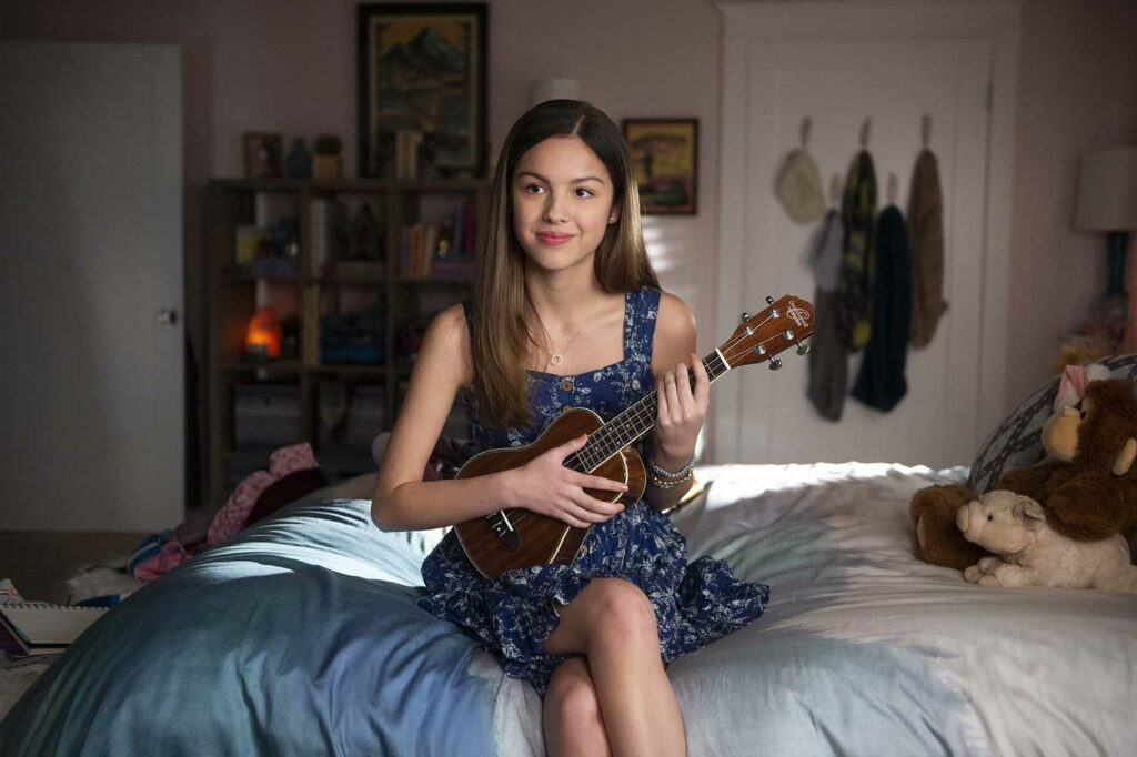 Young Olivia Rodrigo sits on a bed playing a ukulele.