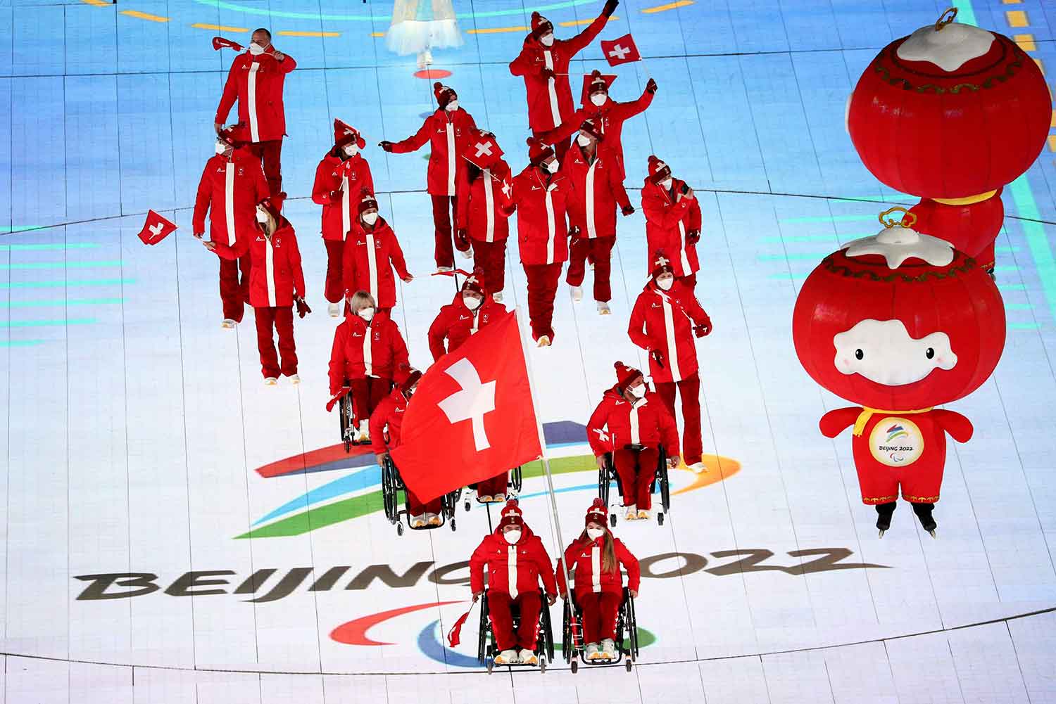Athletes in Swiss uniforms, some with Swiss flags and some using wheelchairs, in a stadium with Beijing 2022 painted on the floor