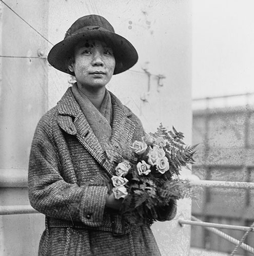 Photo of Mabel Lee standing outdoors in a coat and hat and holding flowers