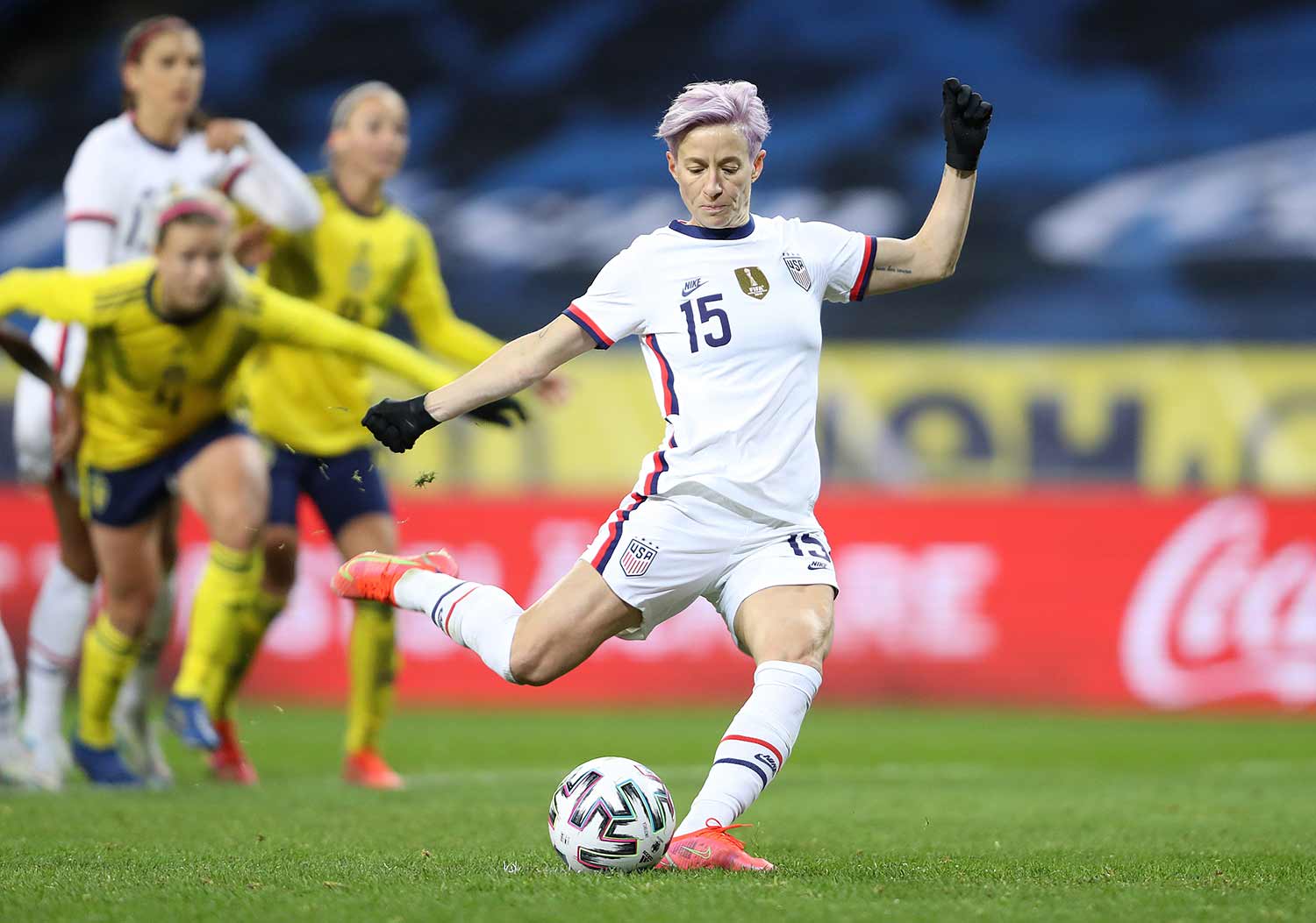 Megan Rapinoe kicking a ball on a soccer field with teammates and members of the opposing team in the background