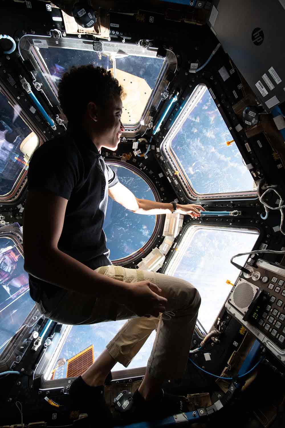 Jessica Watkins sits at a six-sided window looking at a view of Earth with space station instruments in front of her.