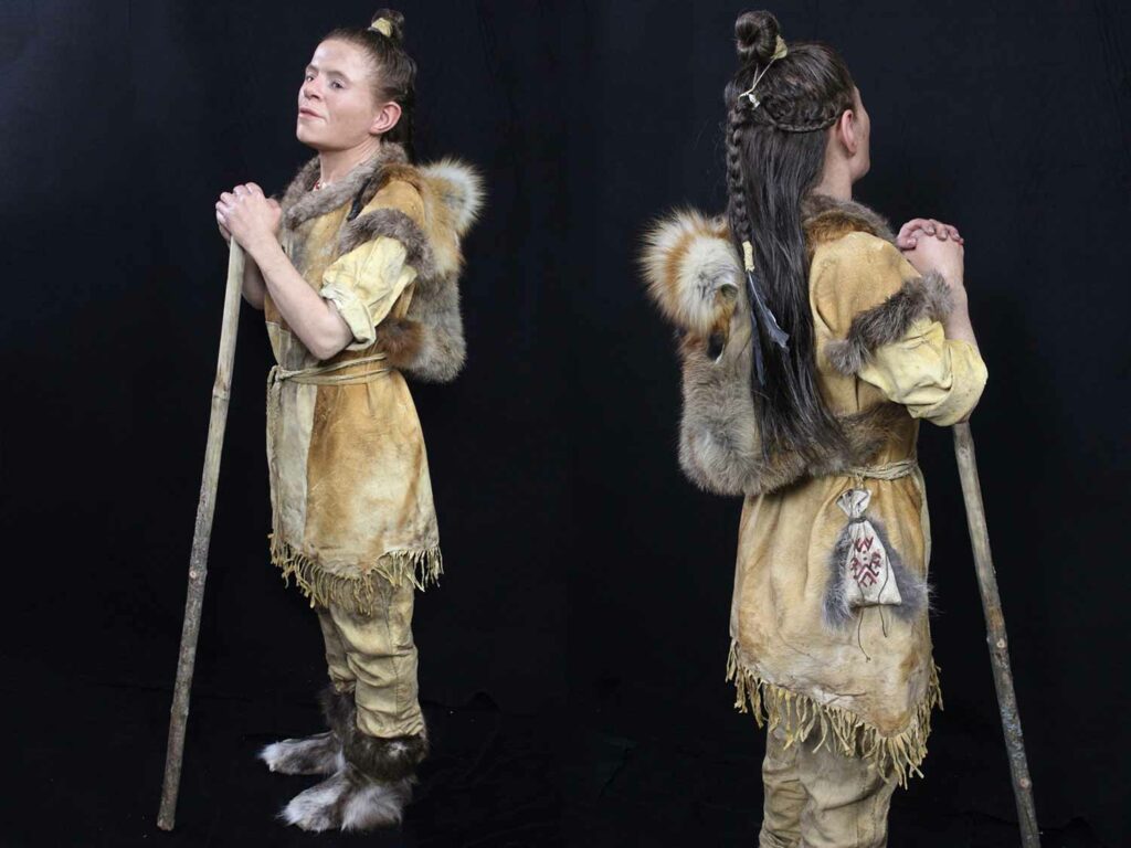 Front and back view of a lifelike model of a woman who lived during the stone age wearing fur and leather and holding a walking stick