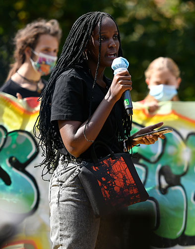 A teen girl stands in front of a banner and protesters and speaks into a microphone.