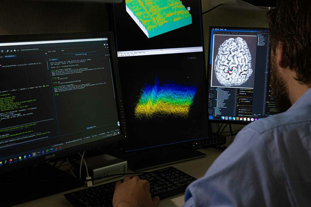 A man sits in front of three keyboard screens showing text, an image of waves, and an image of a brain.