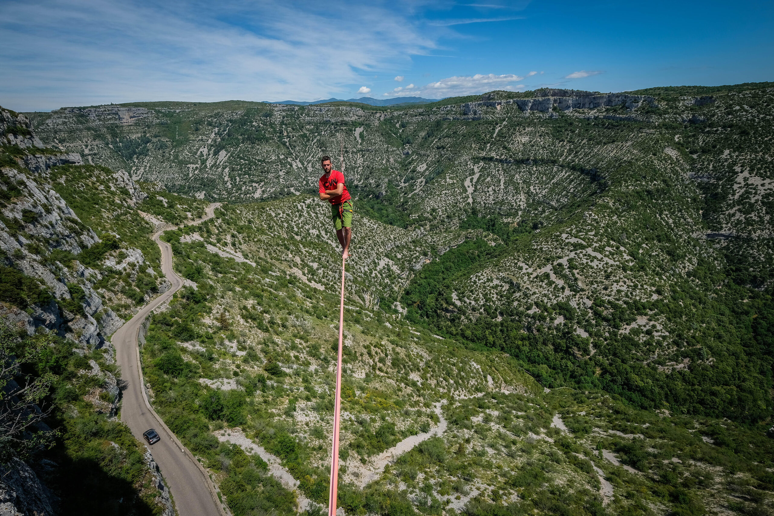 A barefoot man crosses his arms as he walks across a tightrope over road and a green valley.