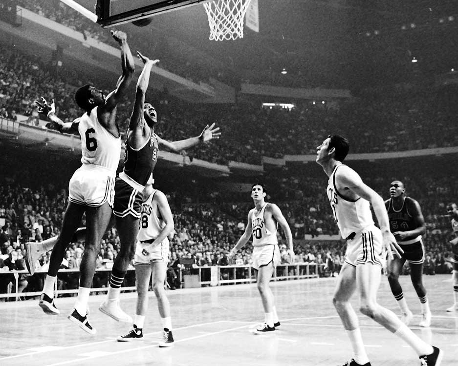 A basketball player tries to block another player’s shot as other players look on.