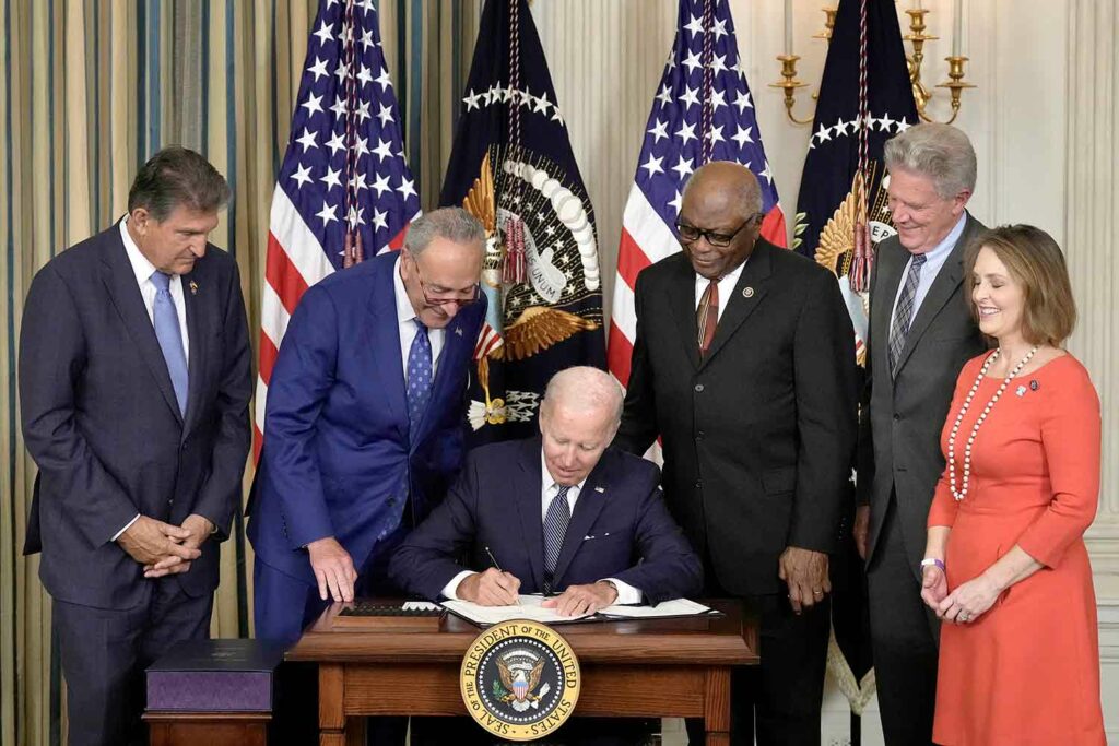 President Biden sits at a table with the presidential seal and signs a paper as Senator Joe Manchin, Senator Chuck Schumer, Representative James Clyburn, Representative Frank Pallone, and Representative Kathy Catson watch.