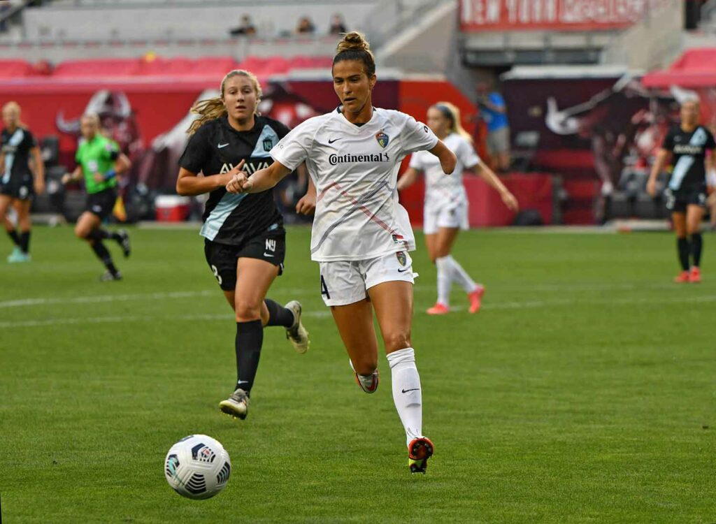 Two women in different uniforms run toward a soccer ball on a field.