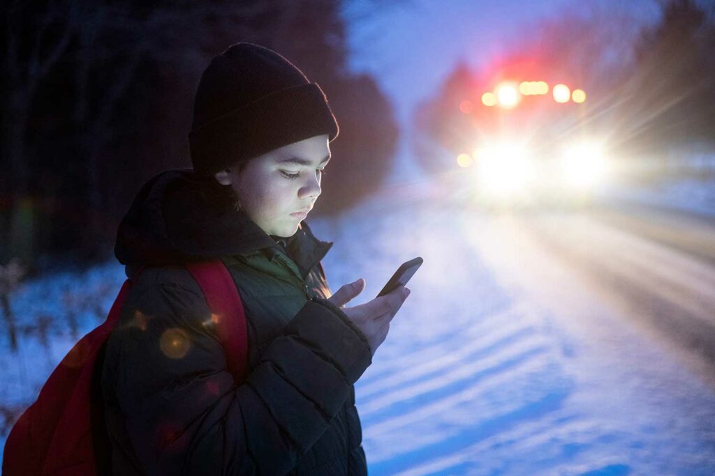 A teen on the side of a snowy road at dawn looks at his phone as a school bus approaches.