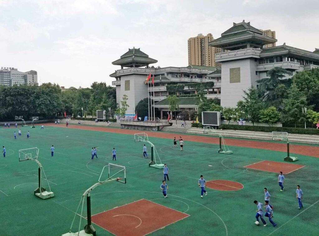 Students kick a ball on a playground next to a school building.