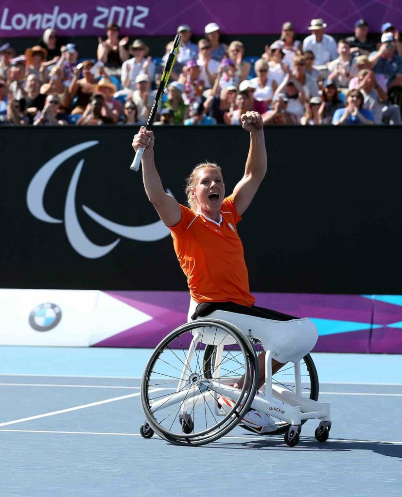 A woman in a wheelchair on a tennis court holds up her arms and smiles as a crowd cheers behind her.