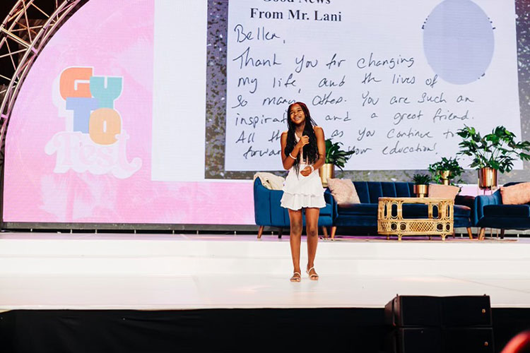 Bellen Woodard stands on a stage holding a microphone in front of an enlarged note that thanks her for being a life-changer.
