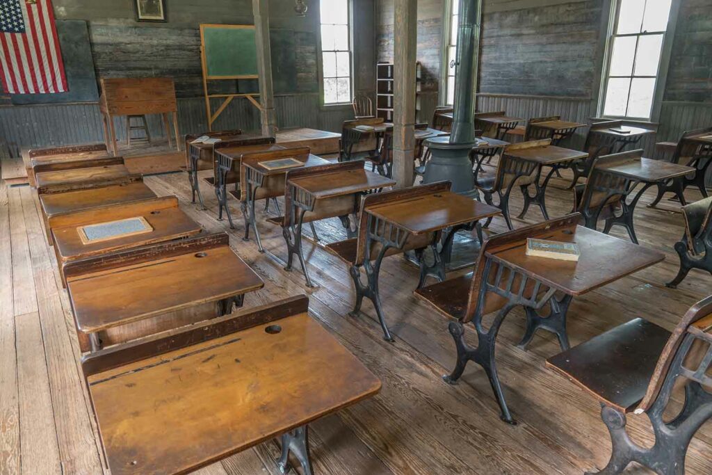 Modern photo of the interior of a one-room schoolhouse with desks, a blackboard, and a wood burning stove.