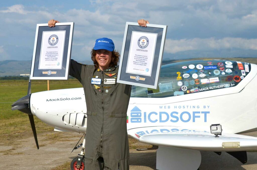A teenage boy in a flight suit smiles as he holds up two framed world record certifications.