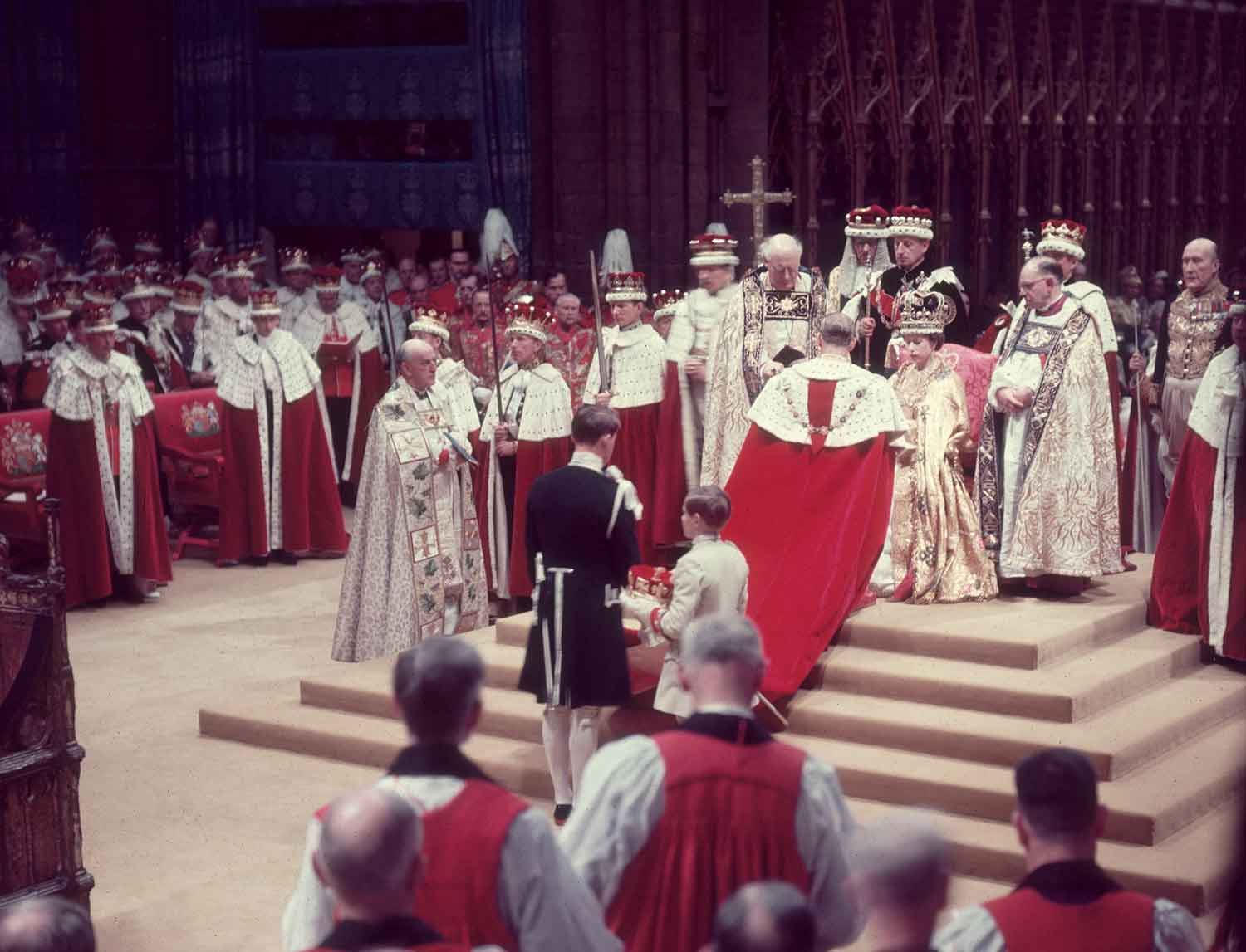 Queen Elizabeth sits on a throne as many religious figures in robes stand before and around her.