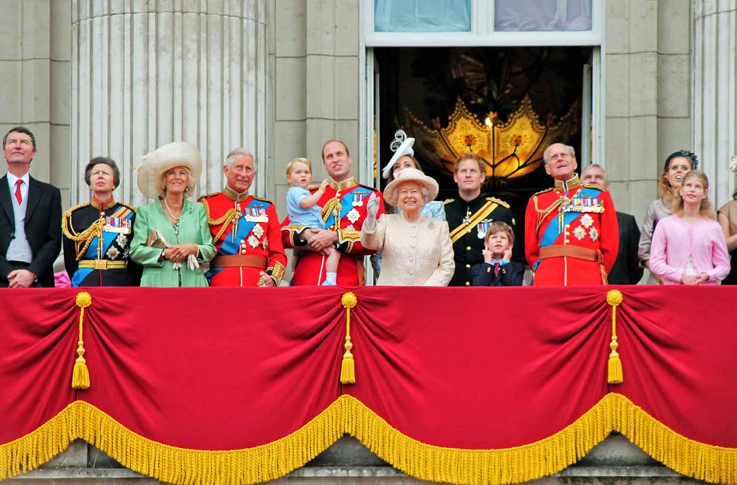 The queen and other members of the royal family are dressed formally as they stand on a balcony and smile.