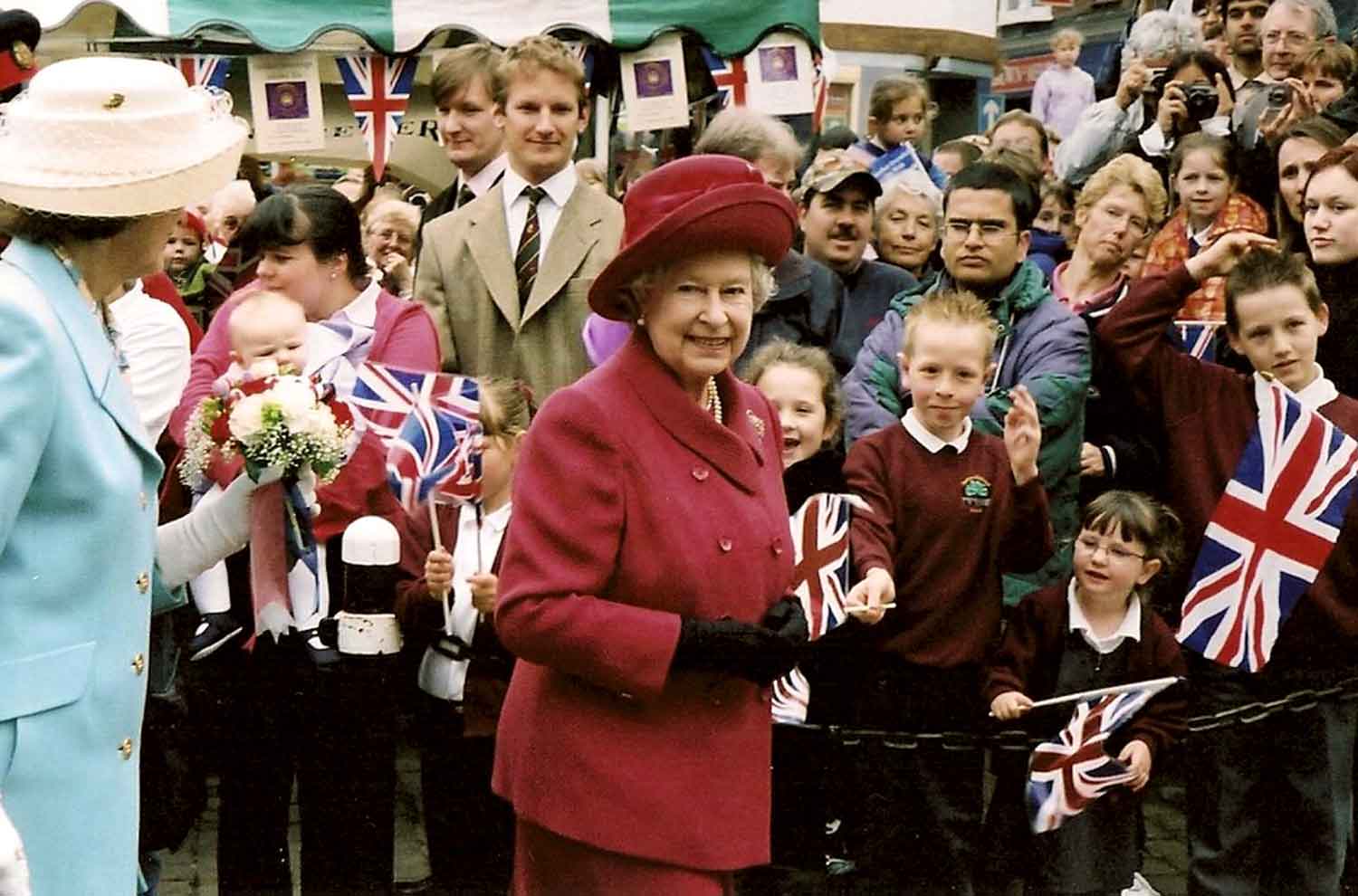 Queen Elizabeth smiles and looks toward the camera as a crowd of people stand behind her, some holding flags of the UK.
