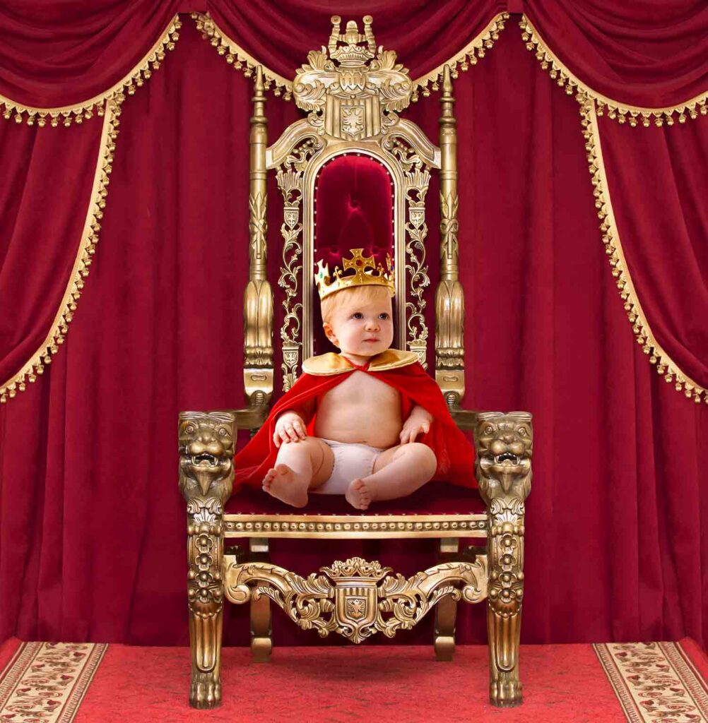 A baby sits on a throne and wears a cape and a crown.