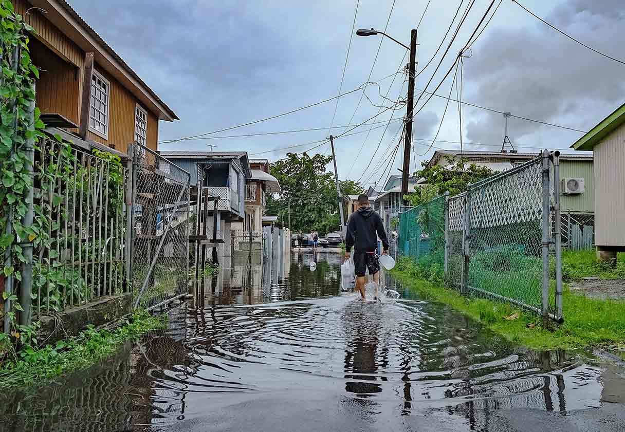 A man carrying a gallon of water walks down a flooded street between rows of houses.