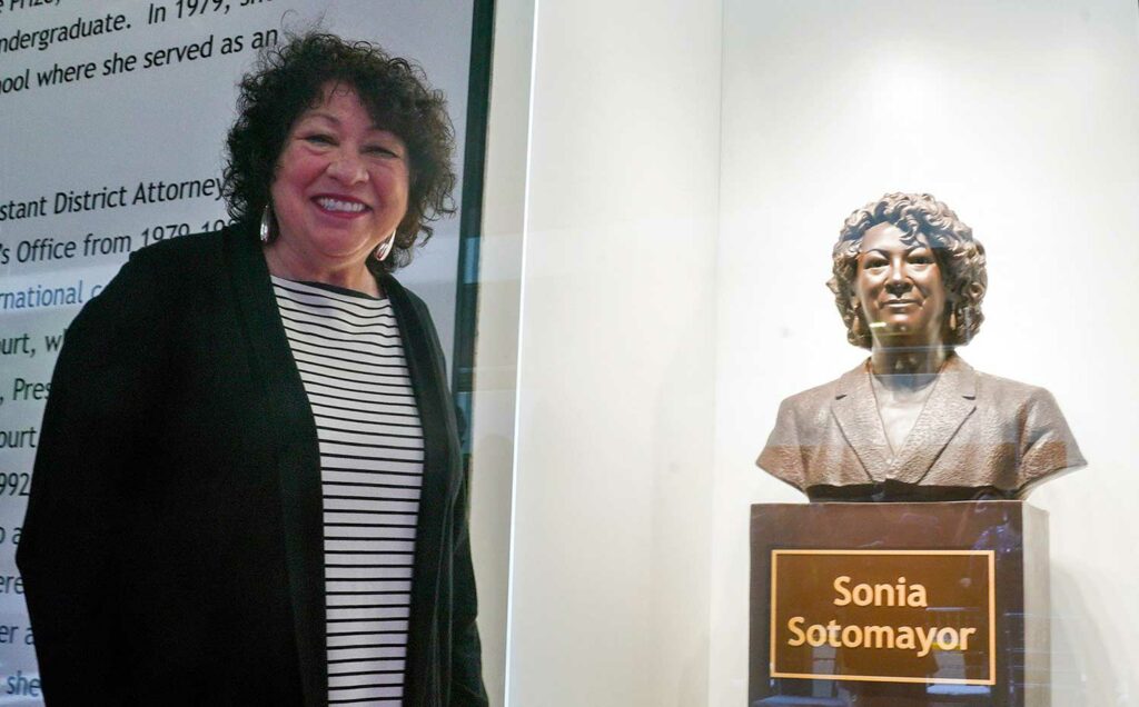 Justice Sonia Sotomayor stands in front of a display case containing a bust of her head.