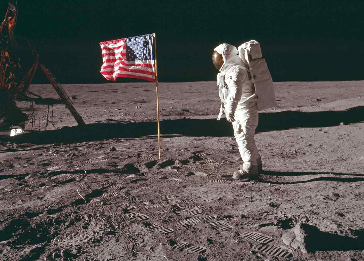A person in a spacesuit stands on the surface of the Moon next to a U.S. flag, which stands upright in the soil.