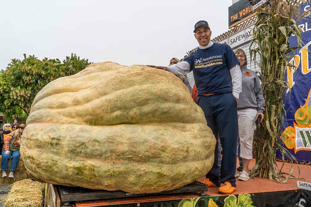 A man stands next to a pumpkin that is almost as large as he is and smiles.