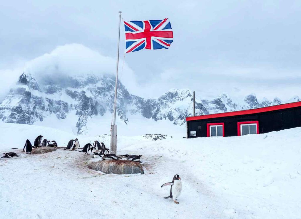 A group of penguins sit or lie around a British flag on an icy landscape with a small building and a mountain in the background.