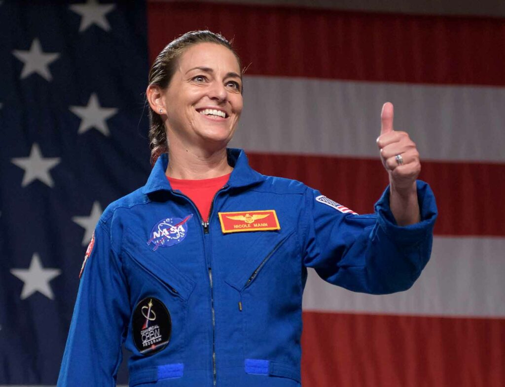 Nicole Mann smiles and gives a thumbs up while wearing a flight suit and standing in front of an American flag.