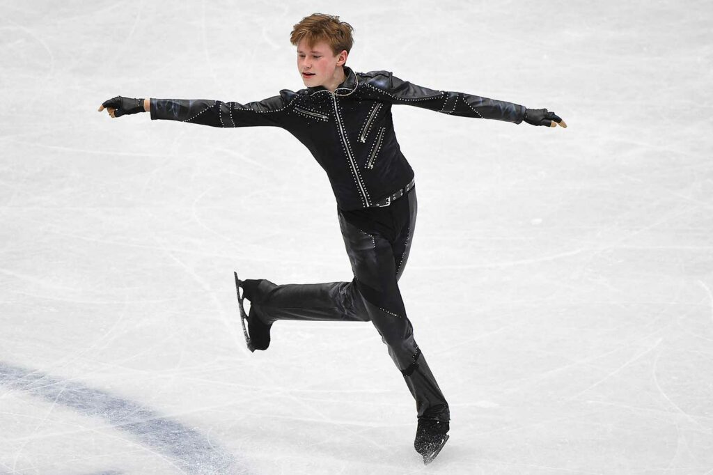 A male figure skater in a black costume and black skates poses on the ice with his arms outstretched.