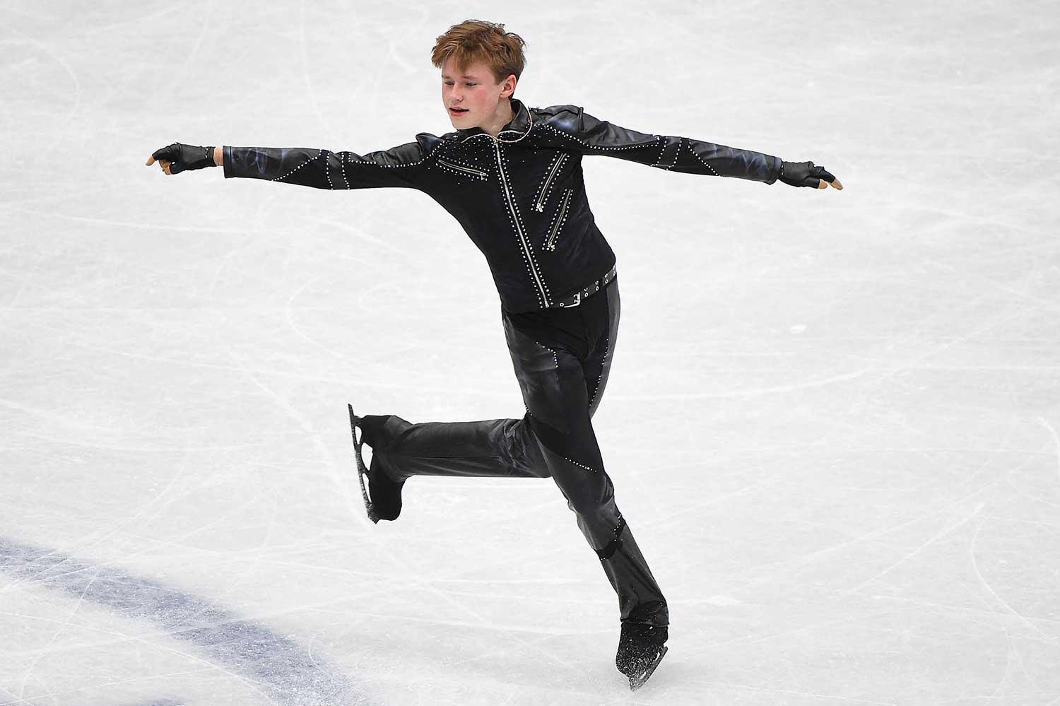 A male figure skater in a black costume and black skates poses on the ice with his arms outstretched.