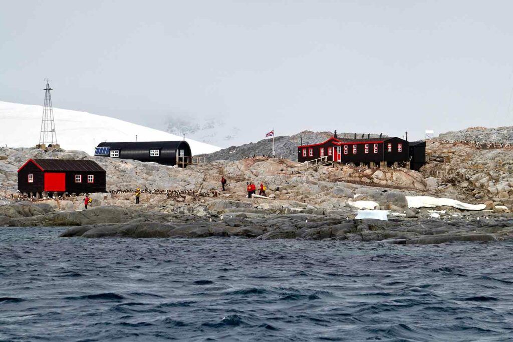 A rocky landscape with three buildings, people, and penguins and a body of water in the foreground.