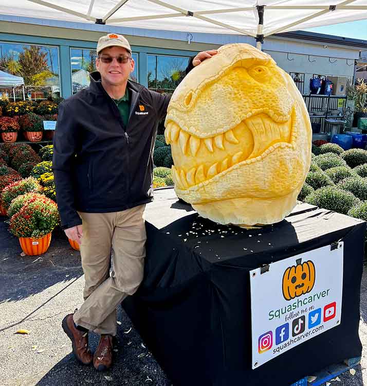 A man stands next to a pumpkin that has been carved to look like the head of a dinosaur.