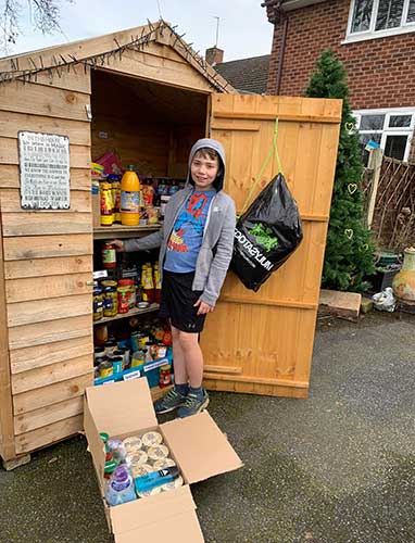 A boy poses in front of a shed that is filled with food. At his feet, a box is filled with supplies.