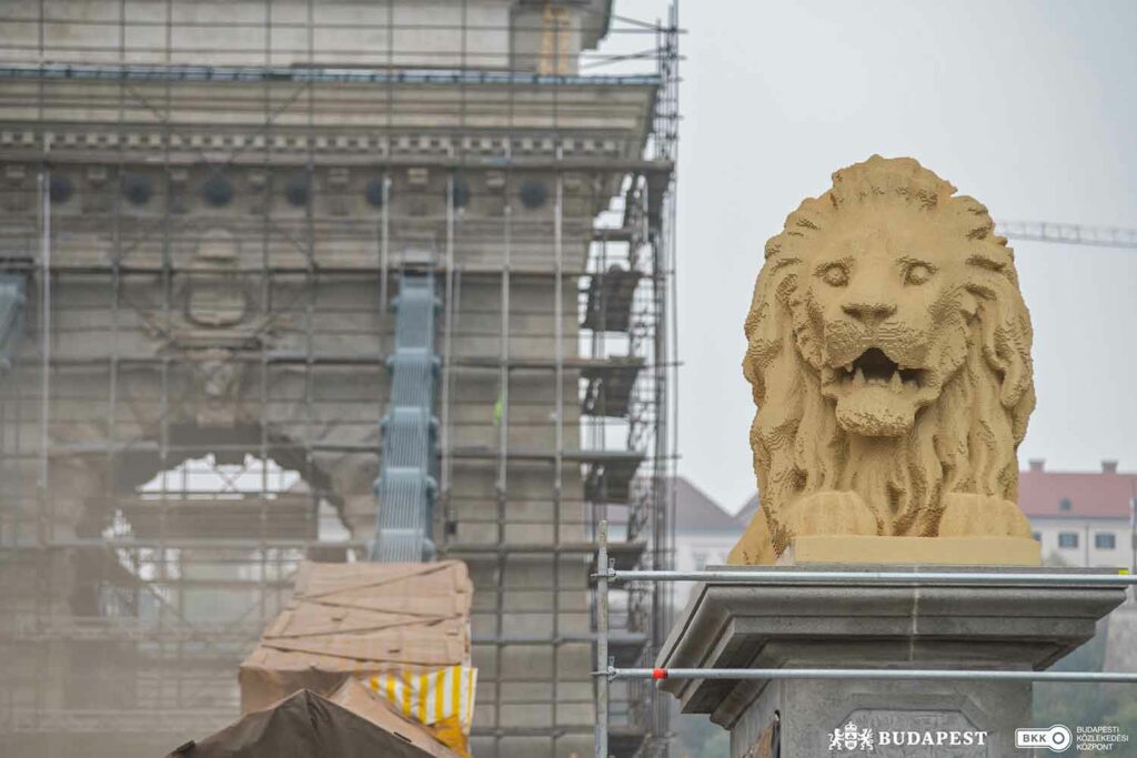 A beige lion made of LEGO bricks lies on a pedestal in front of the entrance to a bridge.