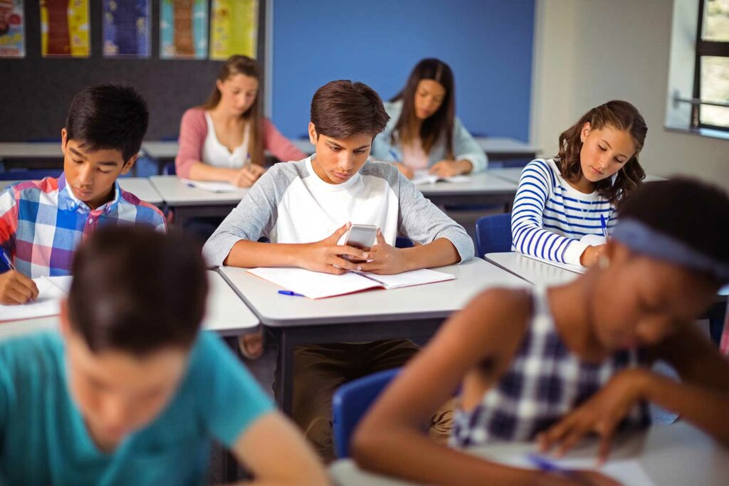 In a classroom, one student looks at his phone while the other students are writing.