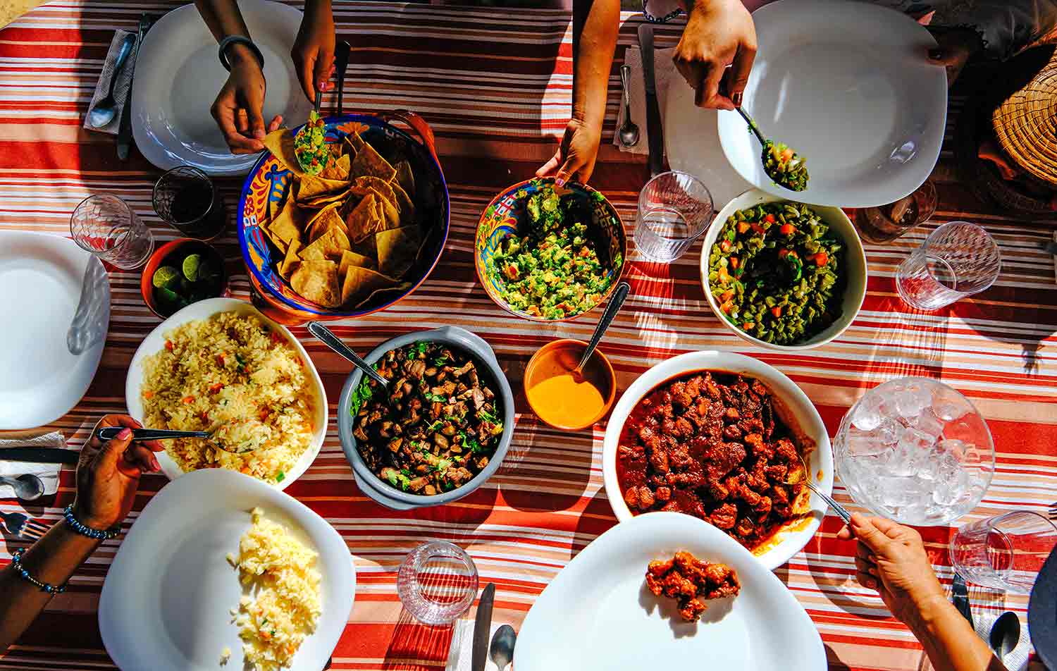 Overhead view of a table with several bowls of food and hands and arms dishing food onto plates.