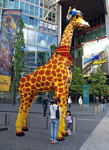 A child poses with a giraffe made of LEGO outside a LEGO store as an adult takes a photo.