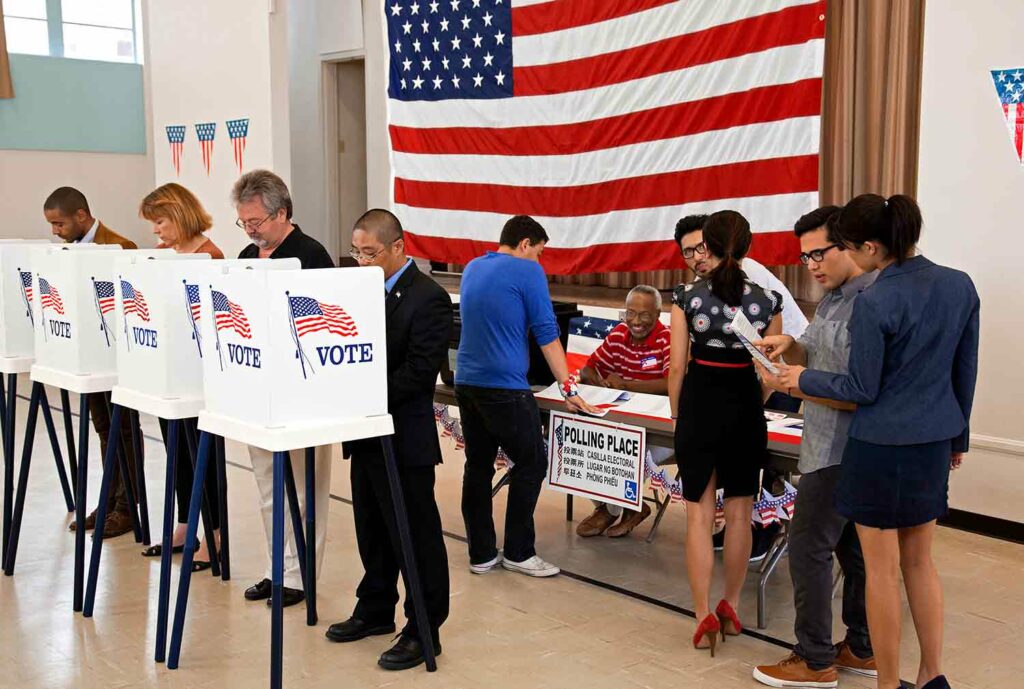 At a voting location, four people stand and cast their votes while others speak to election workers.