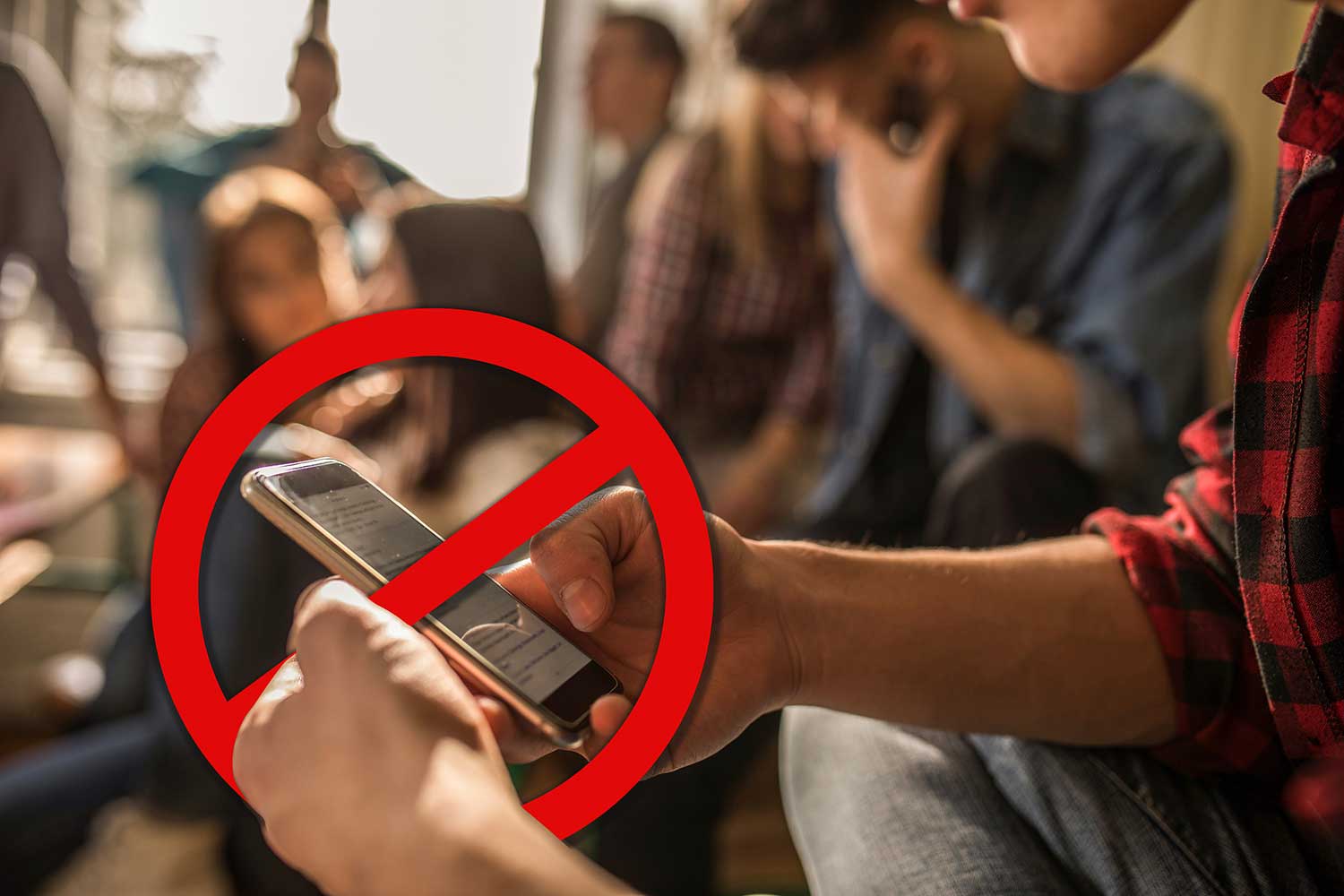 Teen’s hands holding a cell phone with a “no” symbol over it and other teens in background.