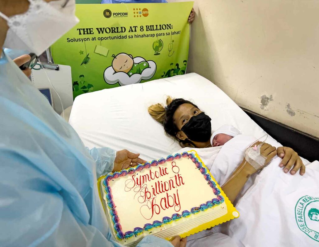 A woman lies in a bed holding a newborn baby as a healthcare worker holds a cake reading Symbolic 8 Billionth Baby