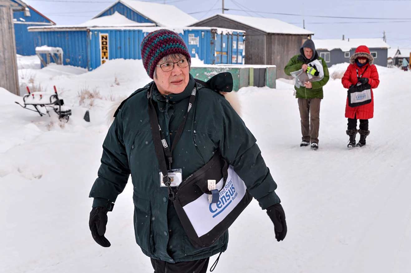 Three people with U.S. Census bags and IDs walk down a snowy road with low lying buildings.