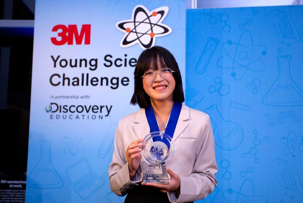 A teen girl smiles and holds up a lucite award in front of a sign reading 3M Young Scientist Challenge.
