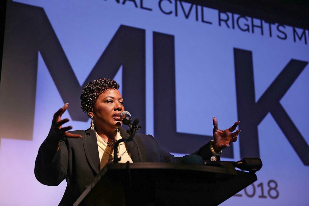 A woman speaks at a podium with a screen behind reading MLK.