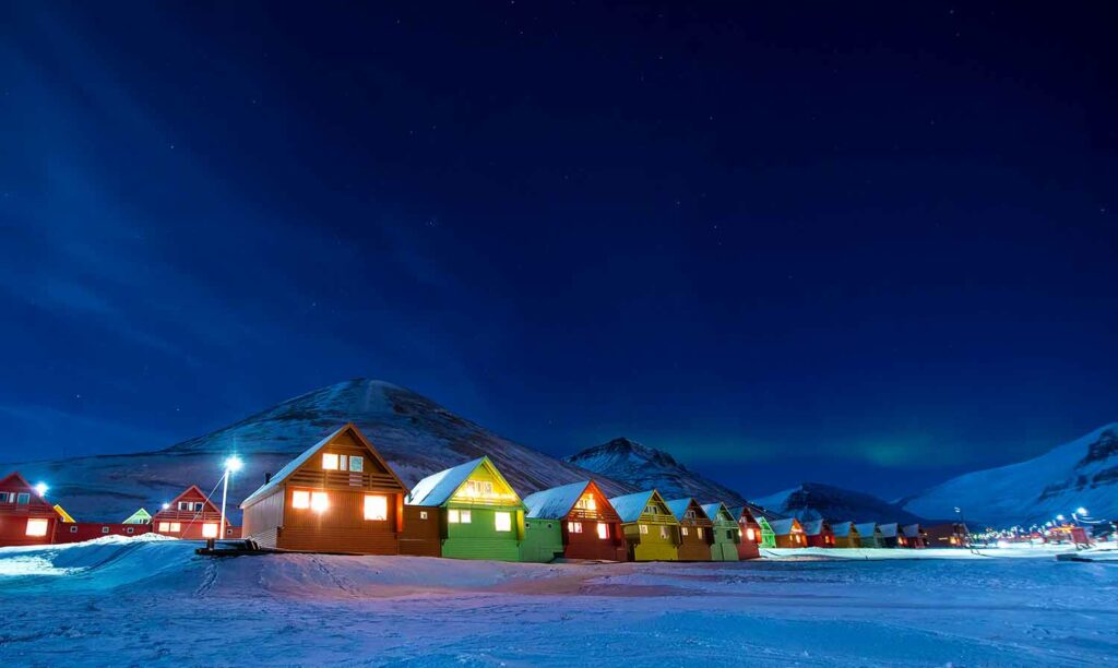 A row of similar houses lit up against a dark sky and a backdrop of snowy mountains.