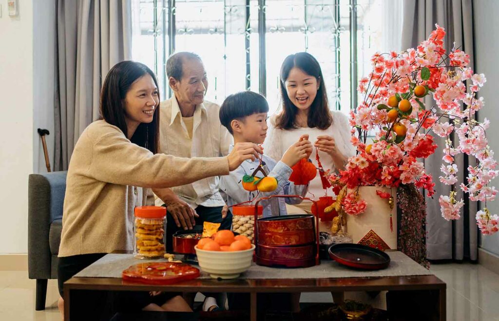 An Asian family of four in a living room decorating a flowered branch with citrus fruit.
