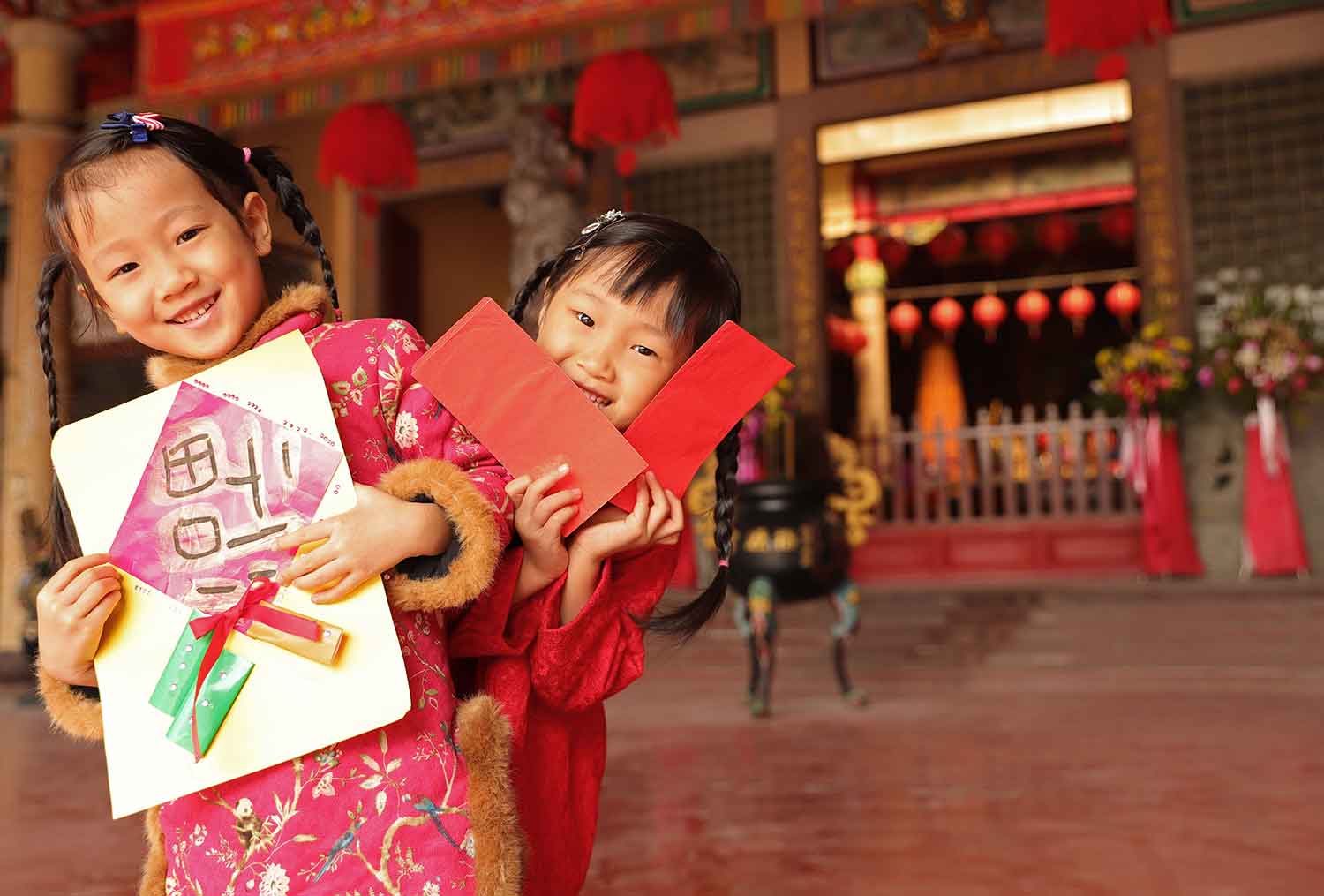 Two smiling girls hold red envelopes and artwork in front of Chinese decorations.