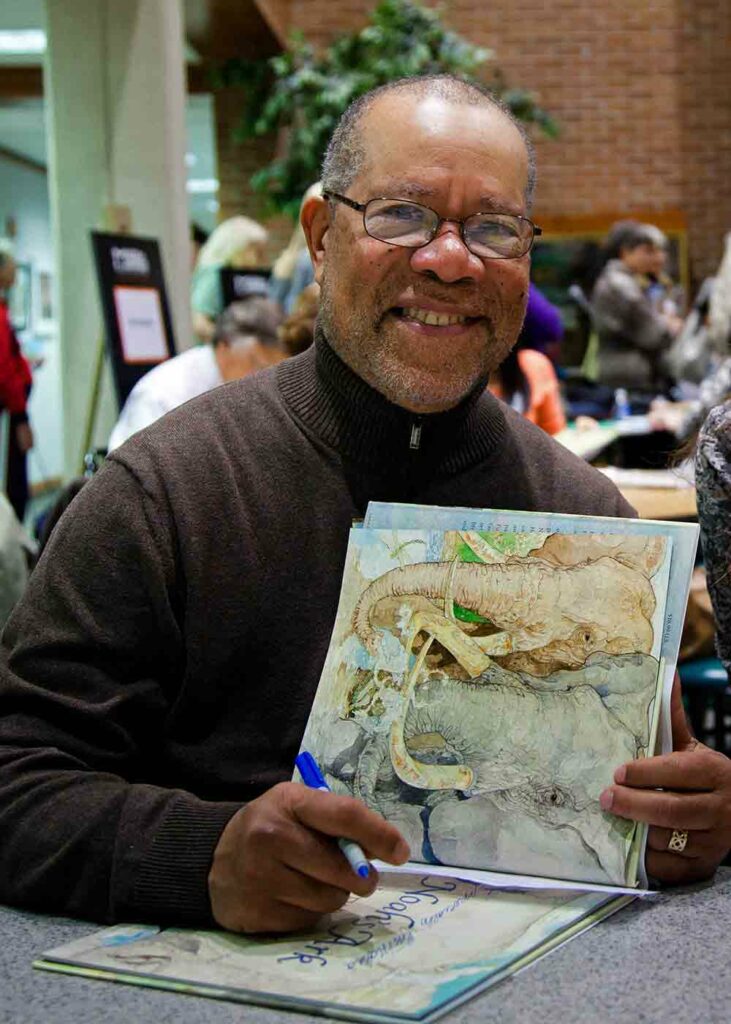 Jerry Pinkney poses with one of his books open holding a pen to sign it.