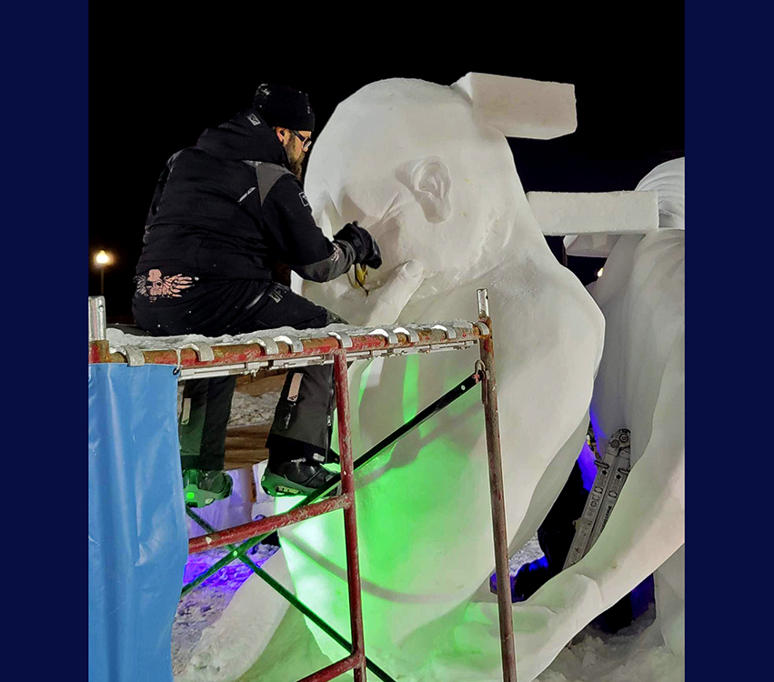 A man sits on scaffolding and carves the figure of a man out of snow.