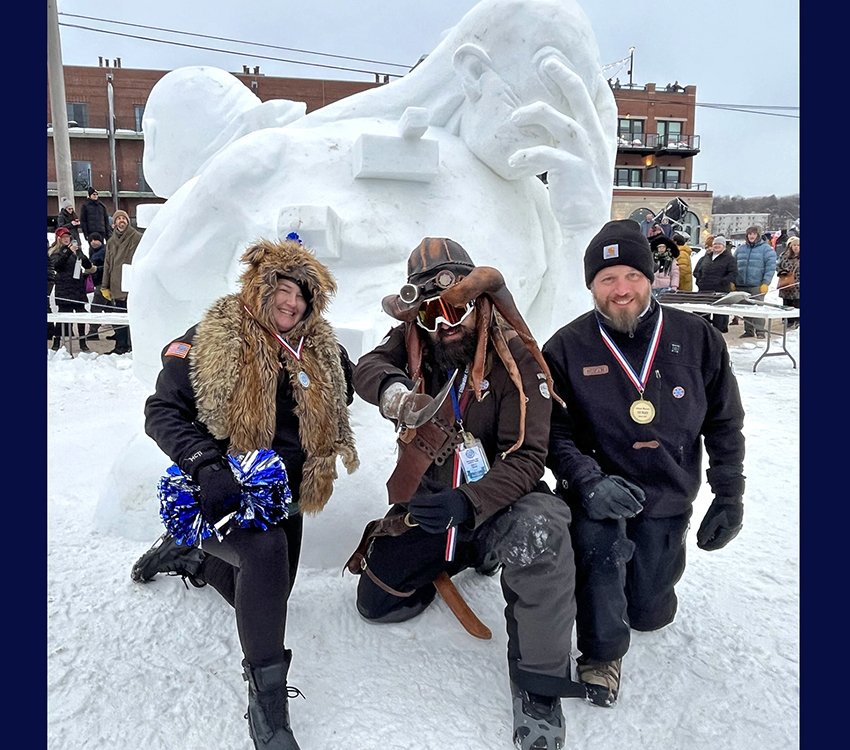 Three people kneel in front of a snow sculpture of a person with his face in his hand.